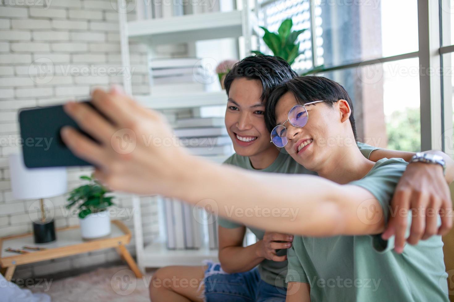 Happy Asian gay couple use mobile phone video call online or using an app on sofa, LGBTQ concept. photo