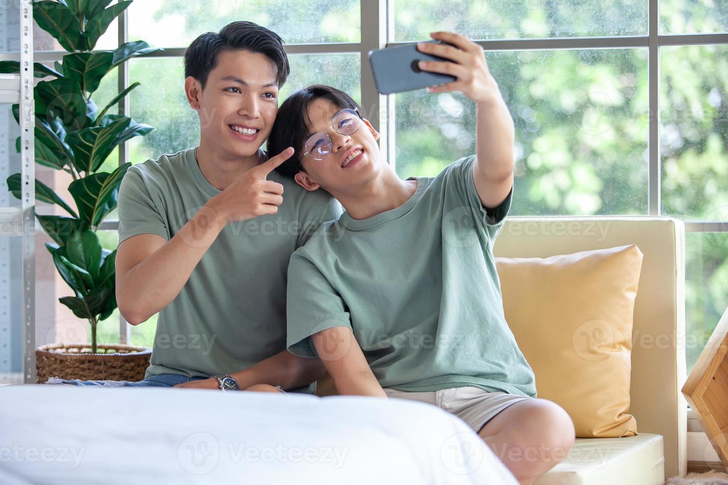 Happy Asian gay couple use mobile phone video call online or using an app on sofa, LGBTQ concept. photo