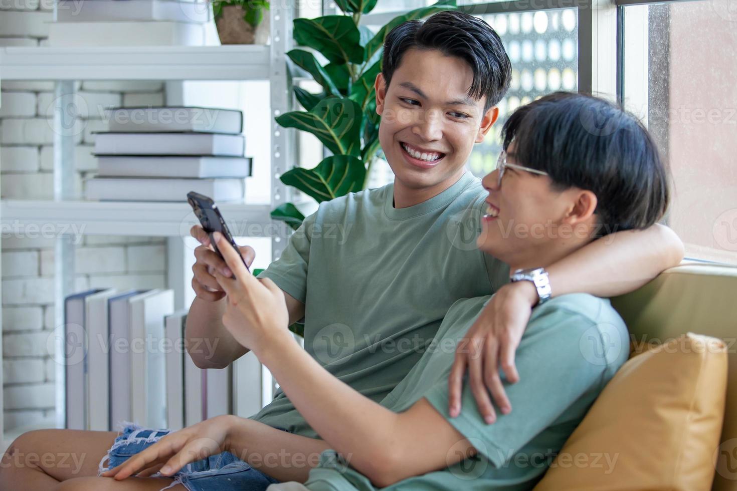Happy Asian gay couple use mobile phone video call online or using an app on sofa, LGBTQ concept. photo