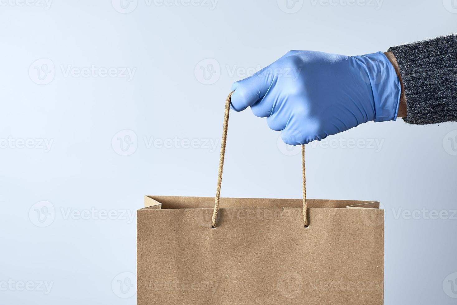 una mano con un guante de goma azul sostiene una bolsa de papel sobre un fondo gris. concepto de compra segura. foto con espacio de copia.