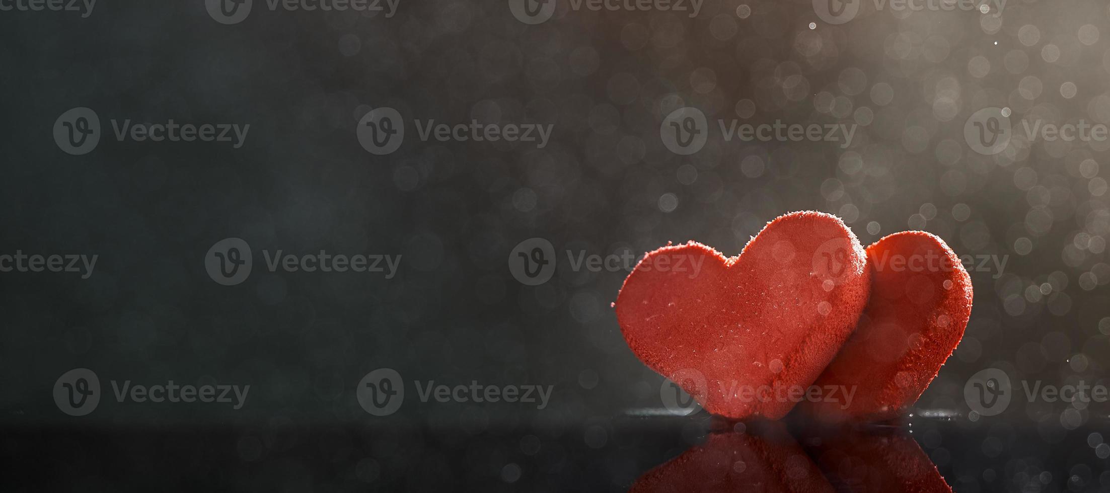 Two red hearts with rain drops illuminated by warm light on dark background with a bokeh. Photo with copy space.