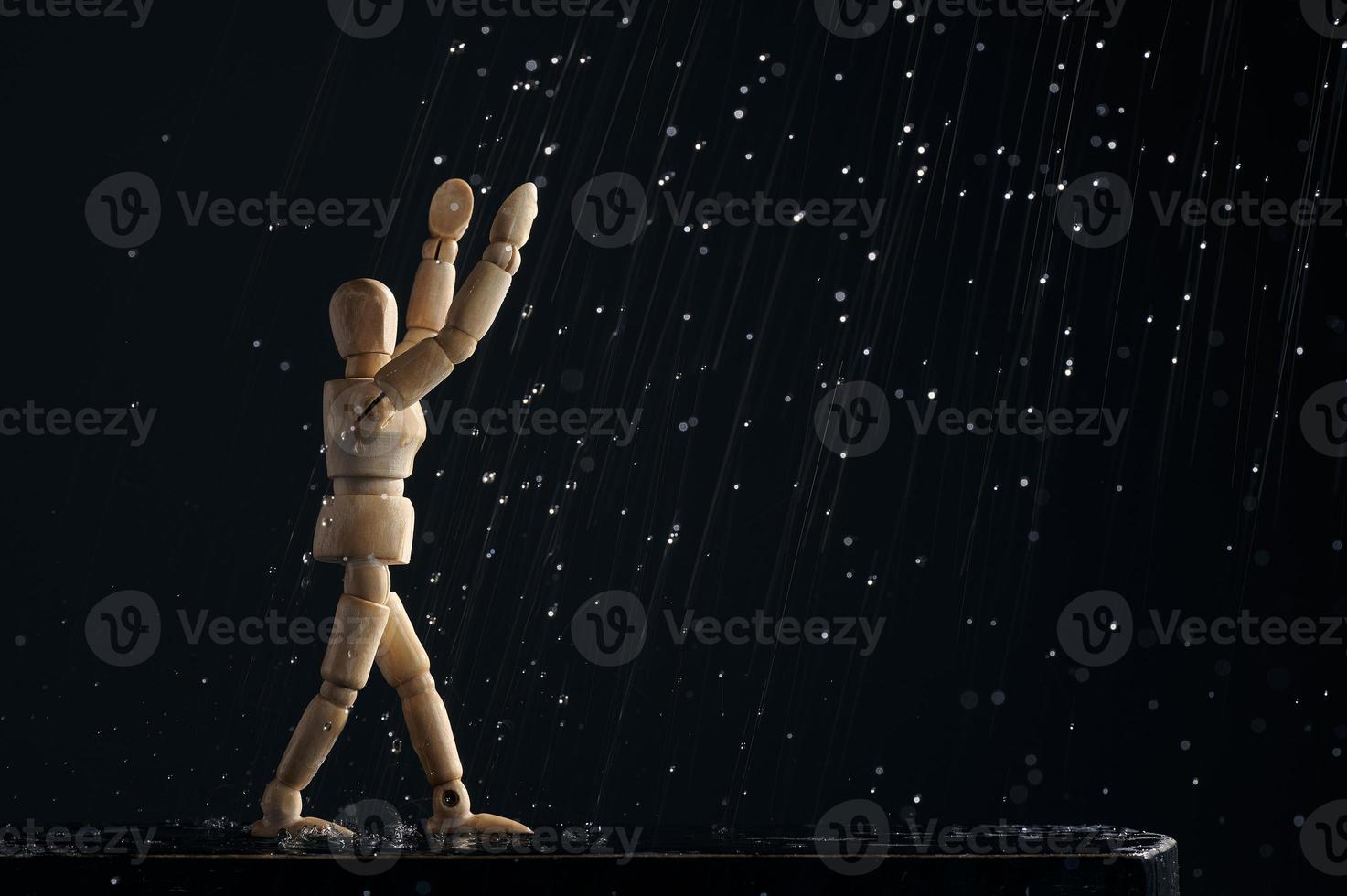 maniquí de madera de un hombre bajo la lluvia levanta las manos sobre un fondo negro. el concepto de alegría. una figura de madera camina a través de charcos. foto