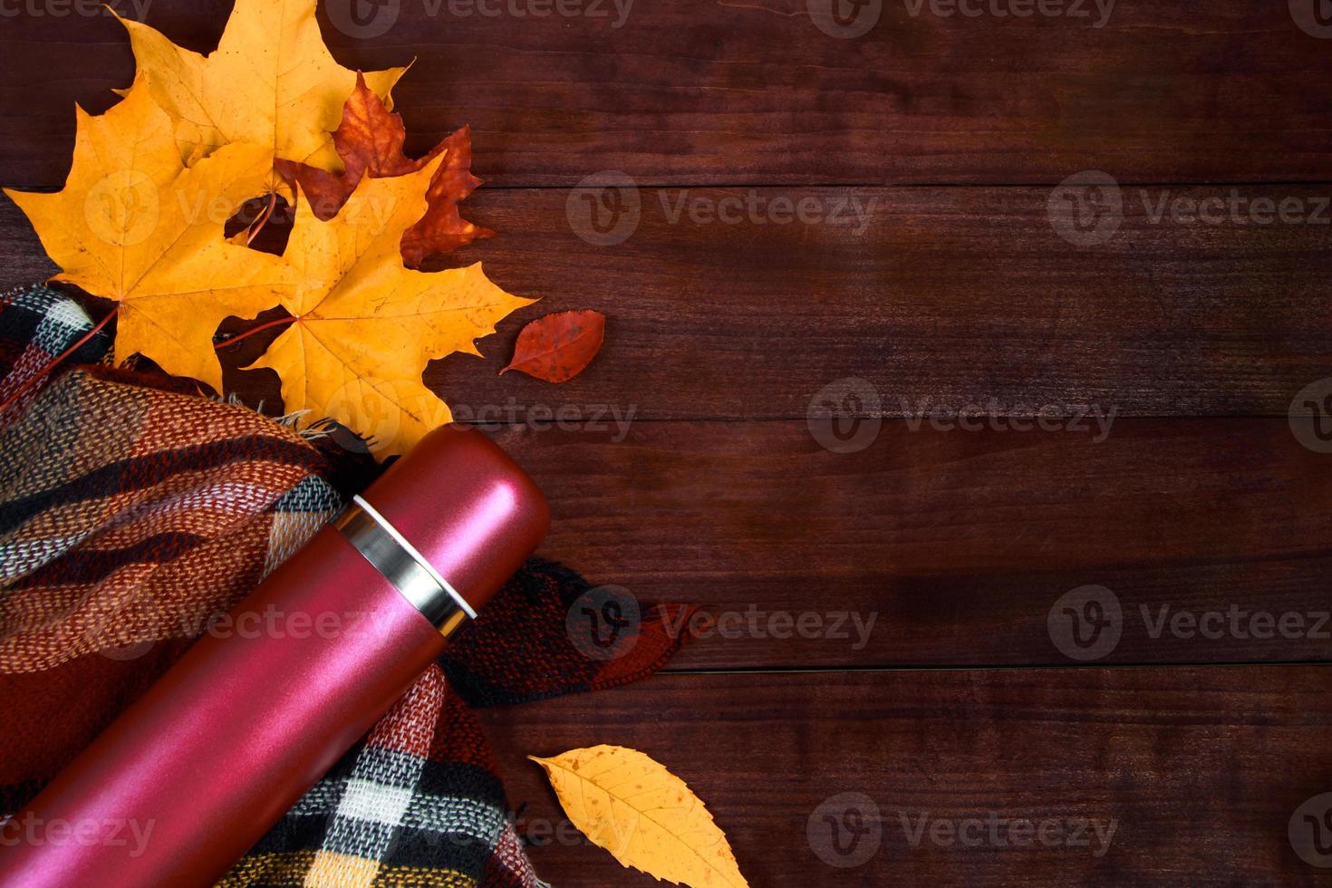 Autumn flat lay. Thermos with hot coffee, plaid and fallen leaves on wooden background. Beginning of the fall season. photo