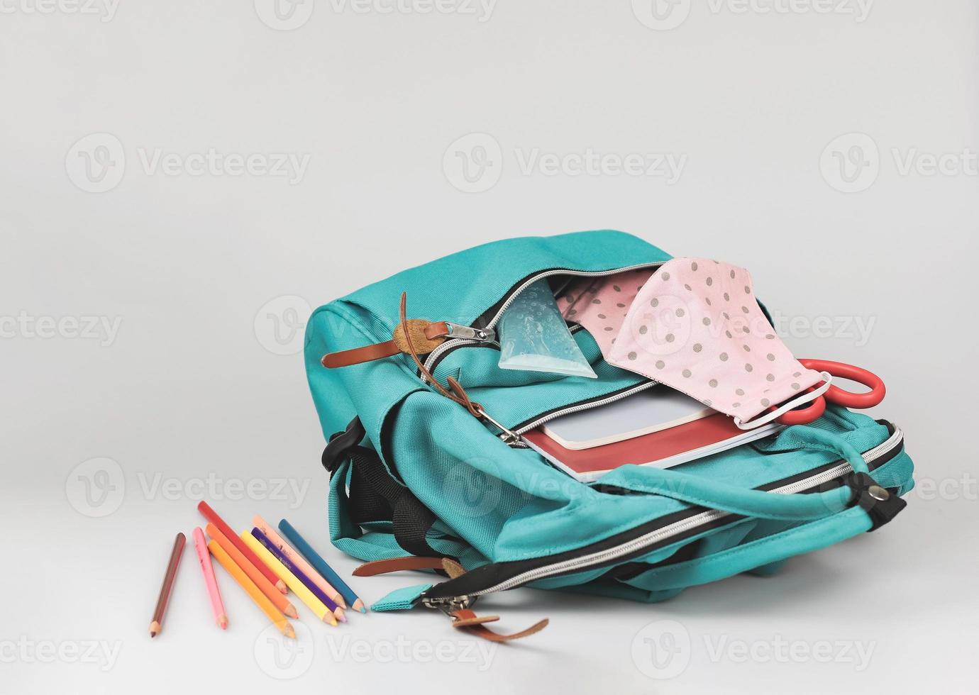back  to school  and new normal  concept.Front view of  pink polka dots fabric mask , sanitizer gel and  backpack with school supplies on white background. photo