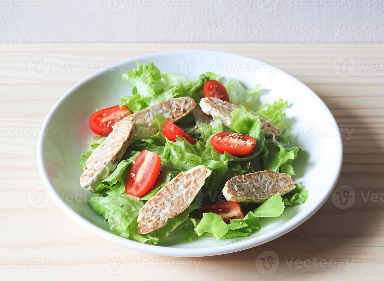 tempeh or tempe salad with tomato and green vegetable in white plate on wooden table. photo