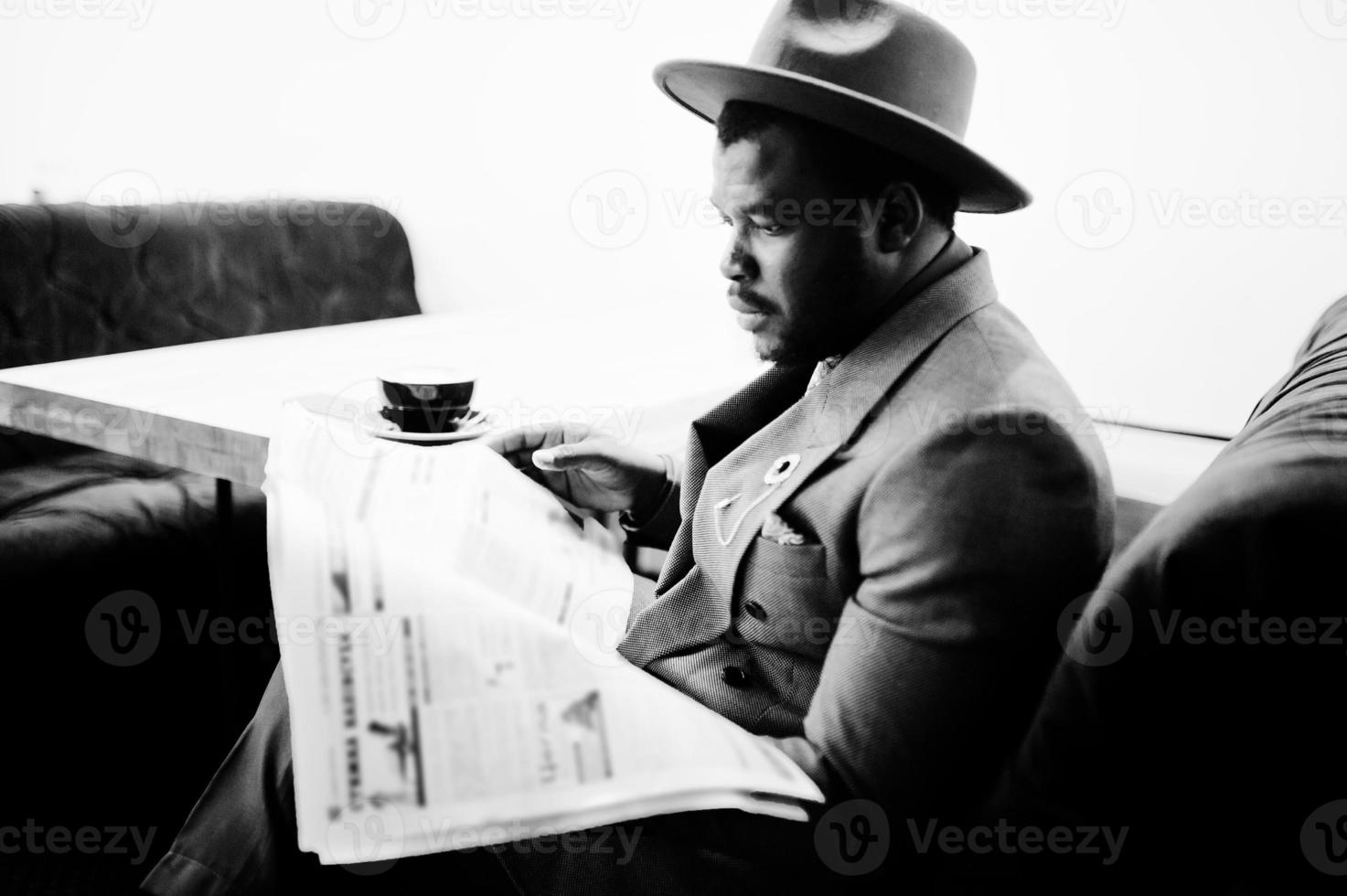 elegante modelo afroamericano con chaqueta gris, corbata y sombrero rojo, bebe café en la cafetería y lee periódicos. foto en blanco y negro.