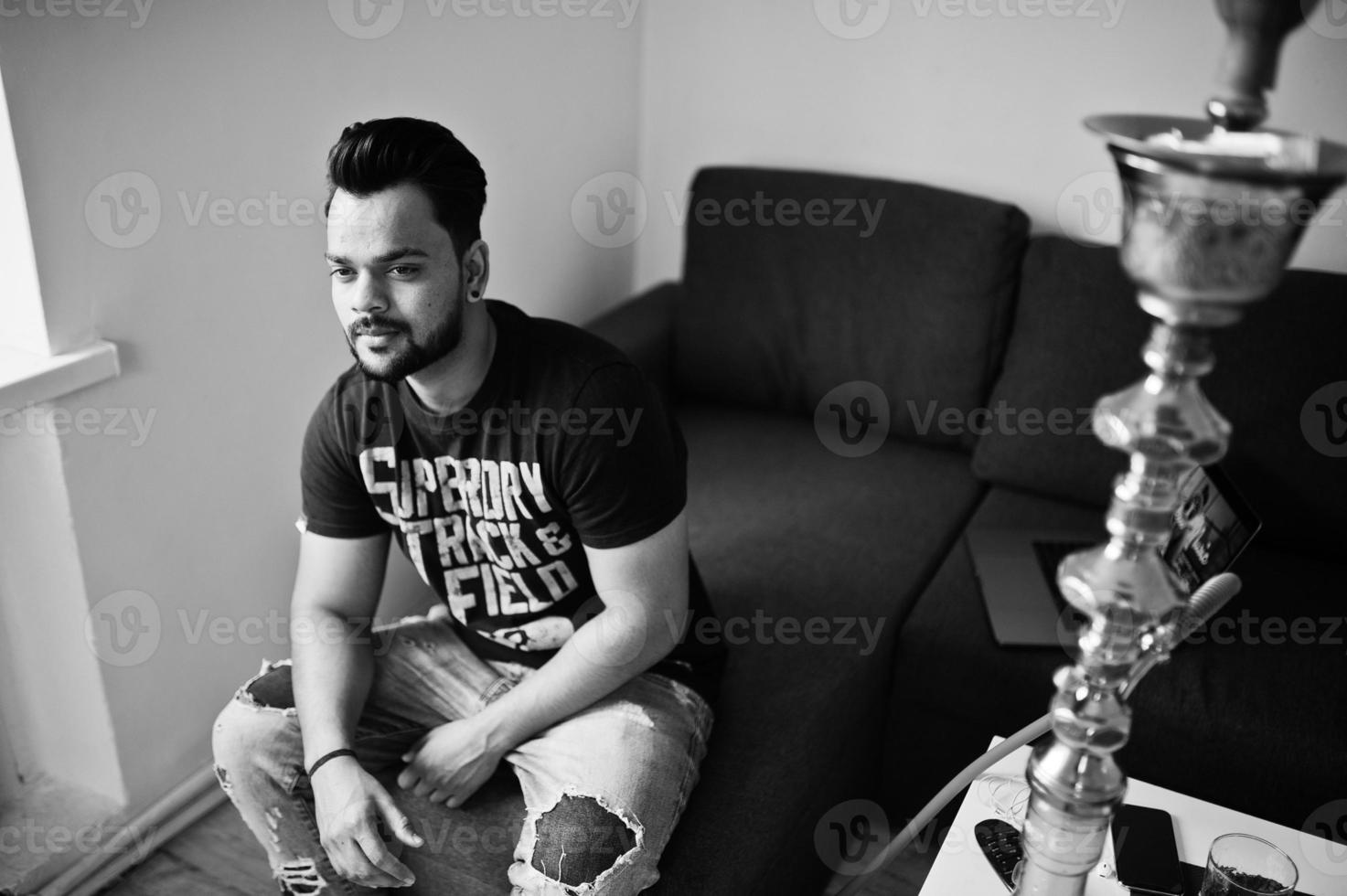 Casual beard indian man sitting at room against hookah. photo