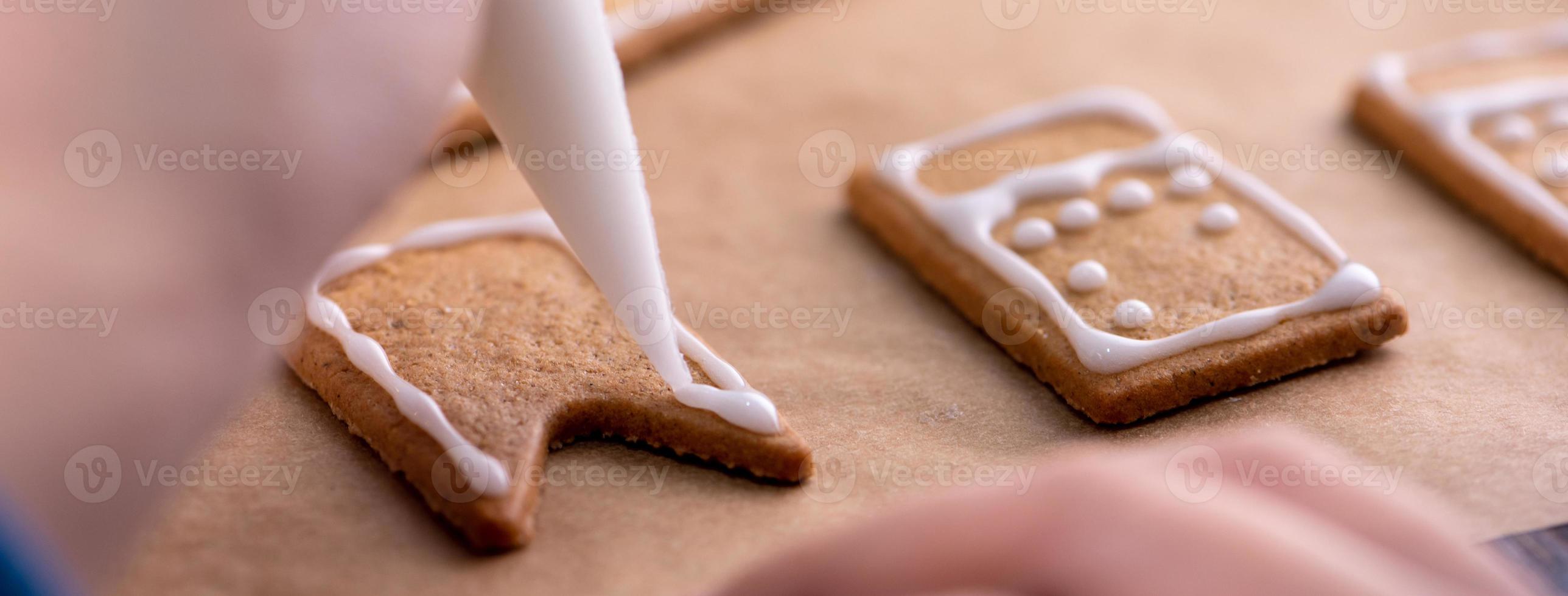 Young woman is decorating Christmas Gingerbread House cookies biscuit at home with frosting topping in icing bag, close up, lifestyle. photo