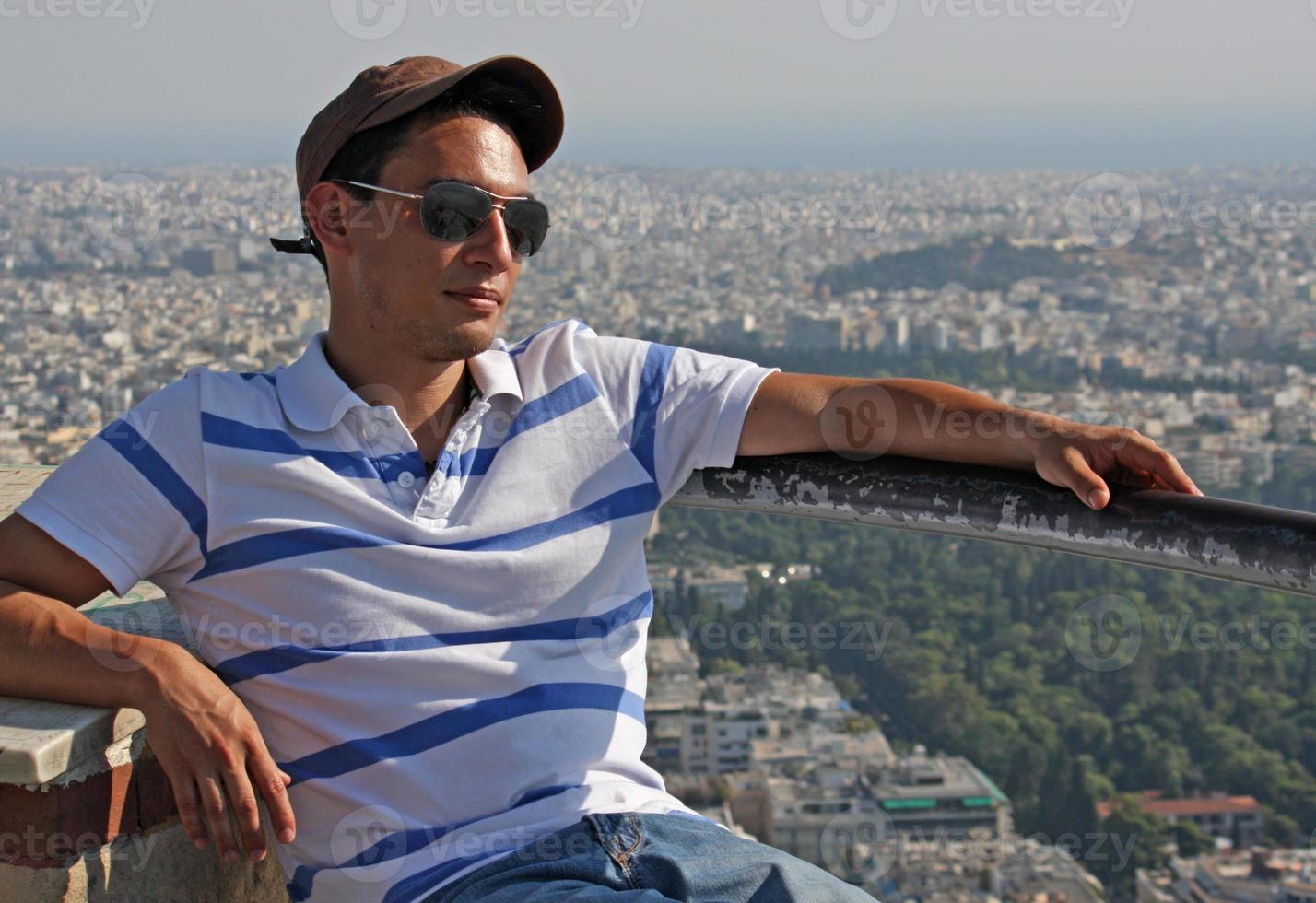 Handsome young man wearing a baseball cap and sun glasses sitting in the sun with the city of Athens in the background photo