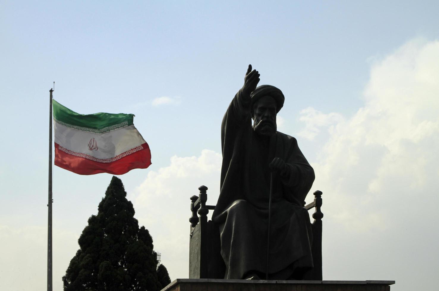 Tehran, Iran - June 10, 2018 - A large Iranian flag waving in the wind behind a statue of Khomeini in Tehran, Iran photo