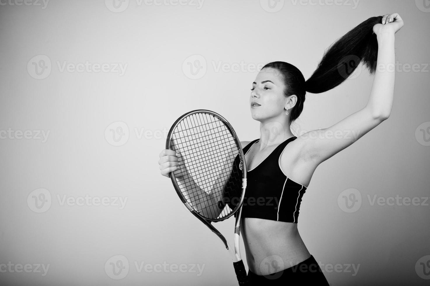 Black and white portrait of beautiful young woman player in sports clothes holding tennis racket while standing against white background. photo