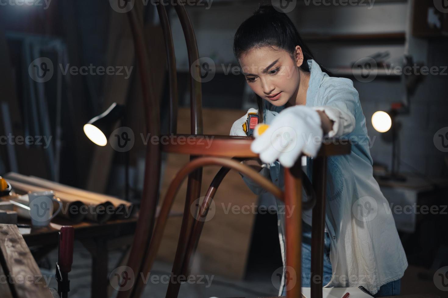 La joven carpintera asiática de diseñador de muebles usa una cinta métrica para medir la silla en el banco de trabajo en el taller de carpintería de madera. foto