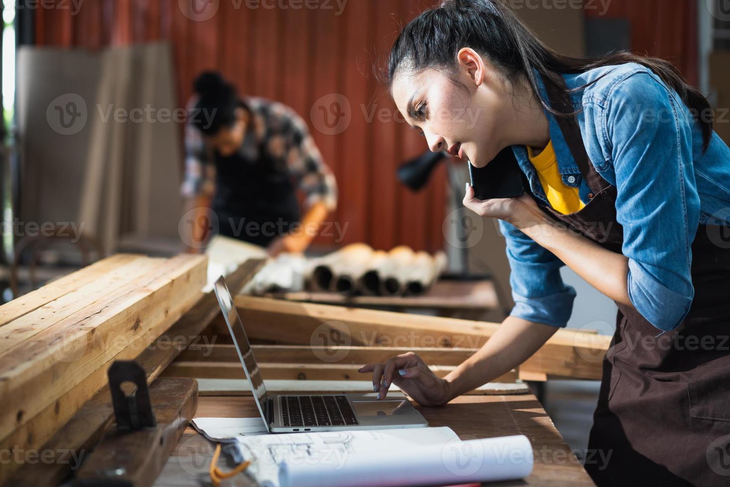 una joven carpintera asiática que usa una computadora portátil mientras habla con un cliente por teléfono en un taller de carpintería de madera. foto