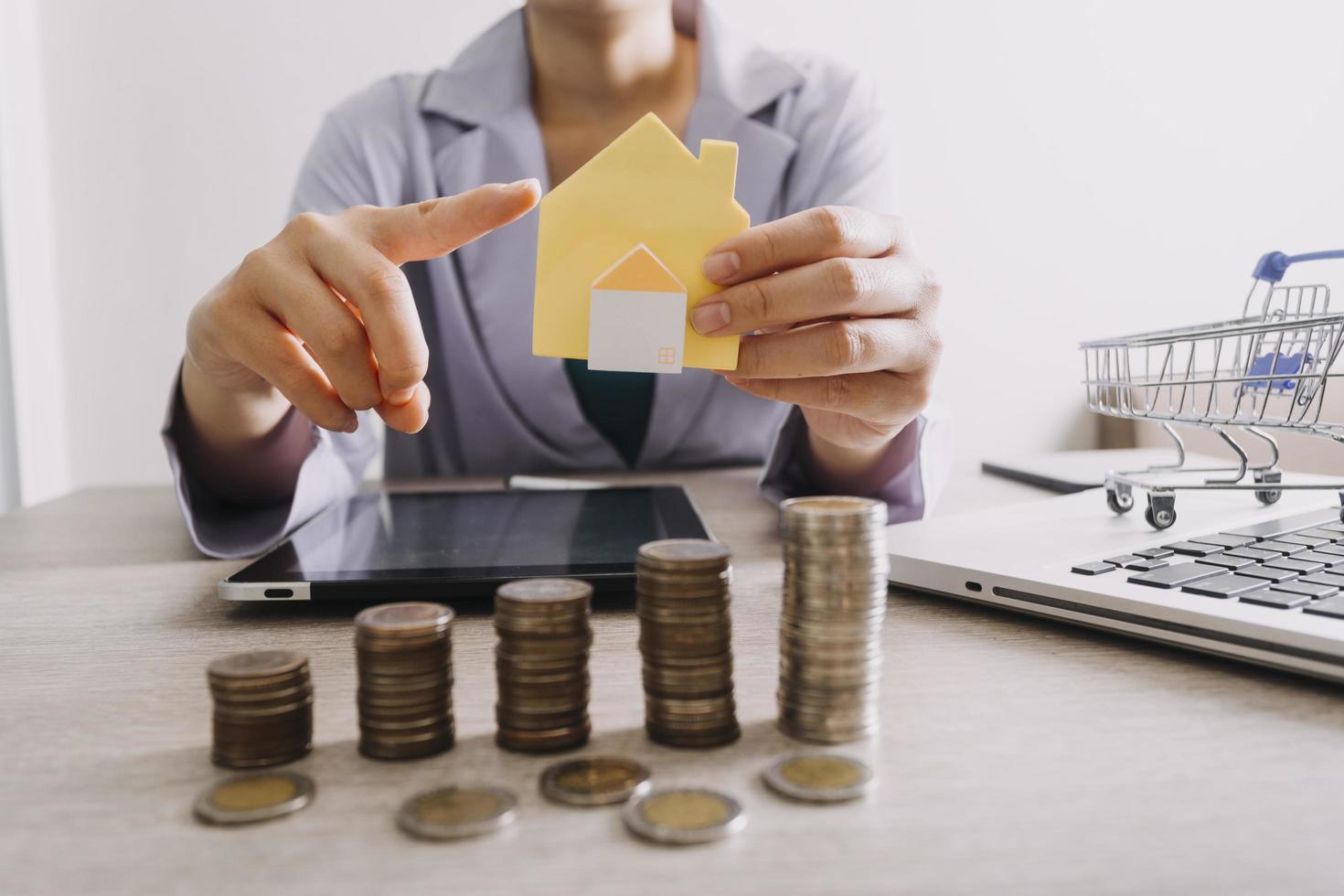 The hand is pressing calculators, piggy bank with wooden house. buy or rent question on note with calculators on desk. Save money and buy house concept. photo