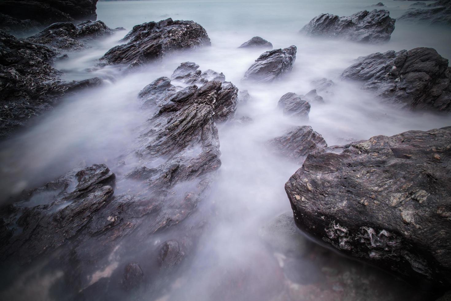 oscuridad de suaves olas del océano al atardecer con piedras en el fondo de la playa. toma de larga exposición del paisaje marino foto