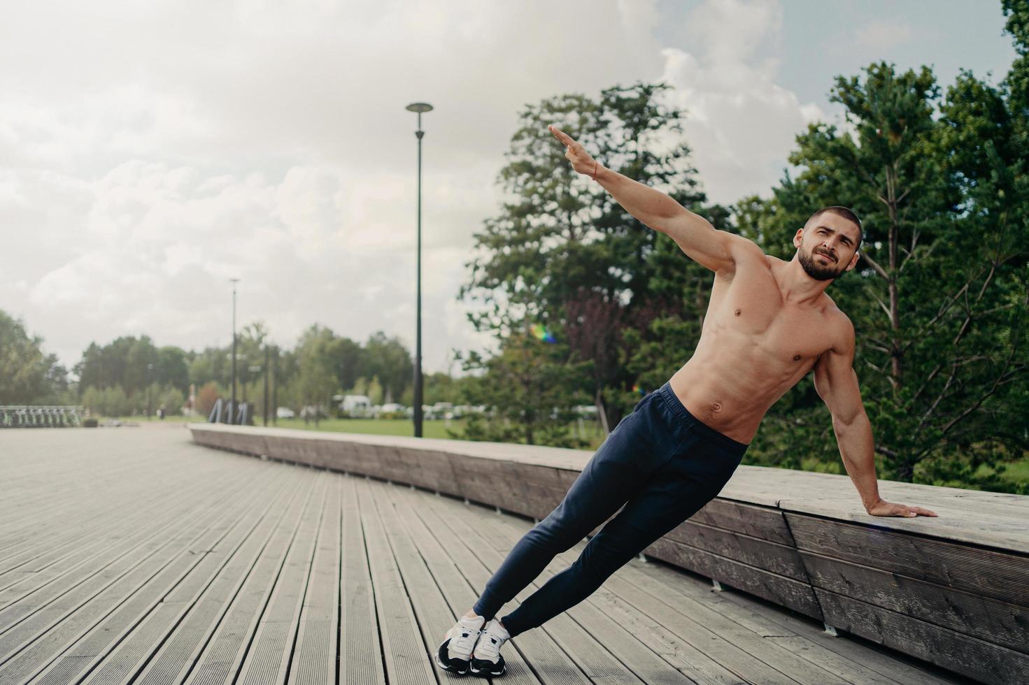 Handsome serious man stands in side plank, has perfect body constitution, poses with naked chest outdoors, keeps arm raised, trains for being healthy, dressed in sport trousers and sneakers. photo