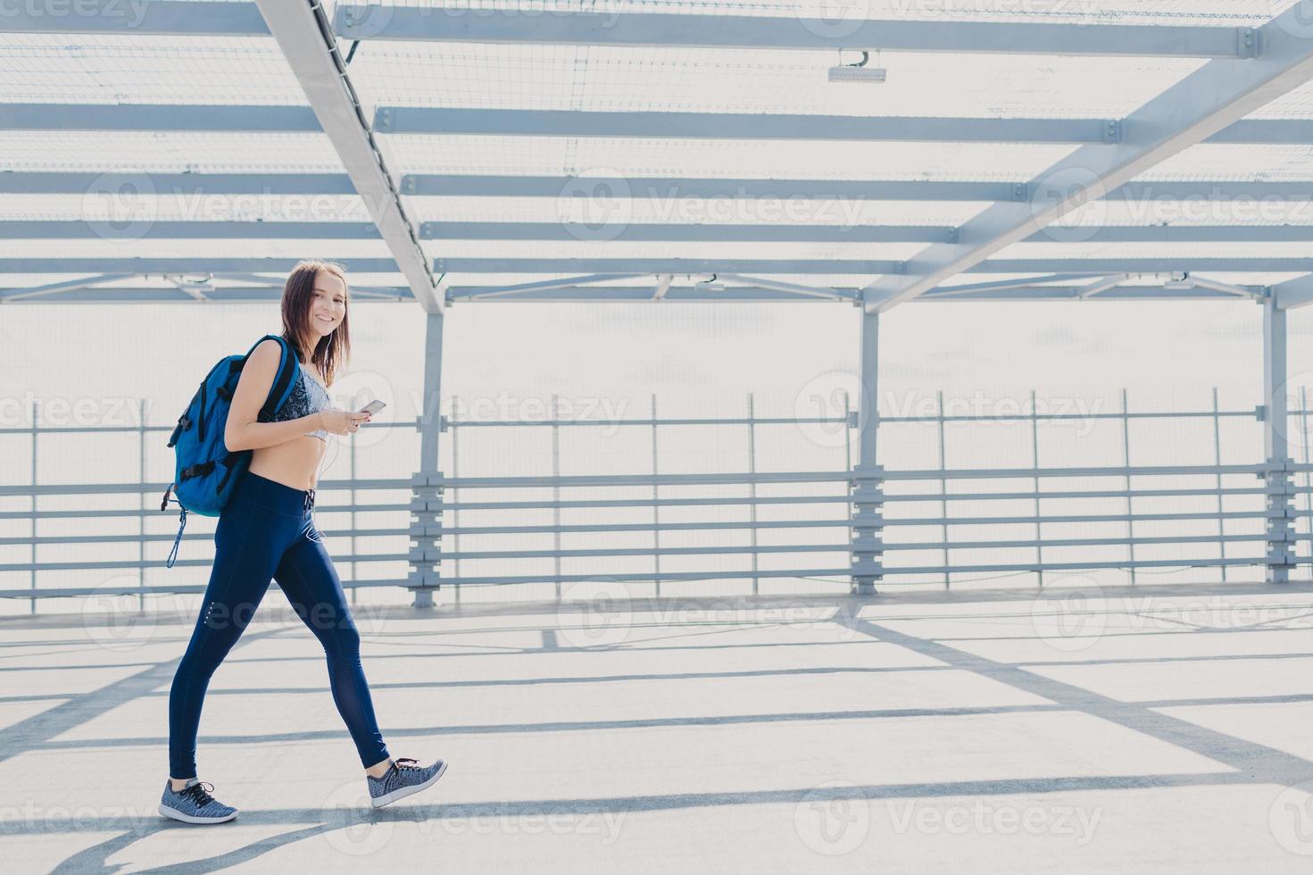 Active female fitness trainer dressed in sportsclothes, sneakers, carries rucksack, uses mobile phone for checking calories, listens music with earphones, uses free wifi connection. People and sport photo