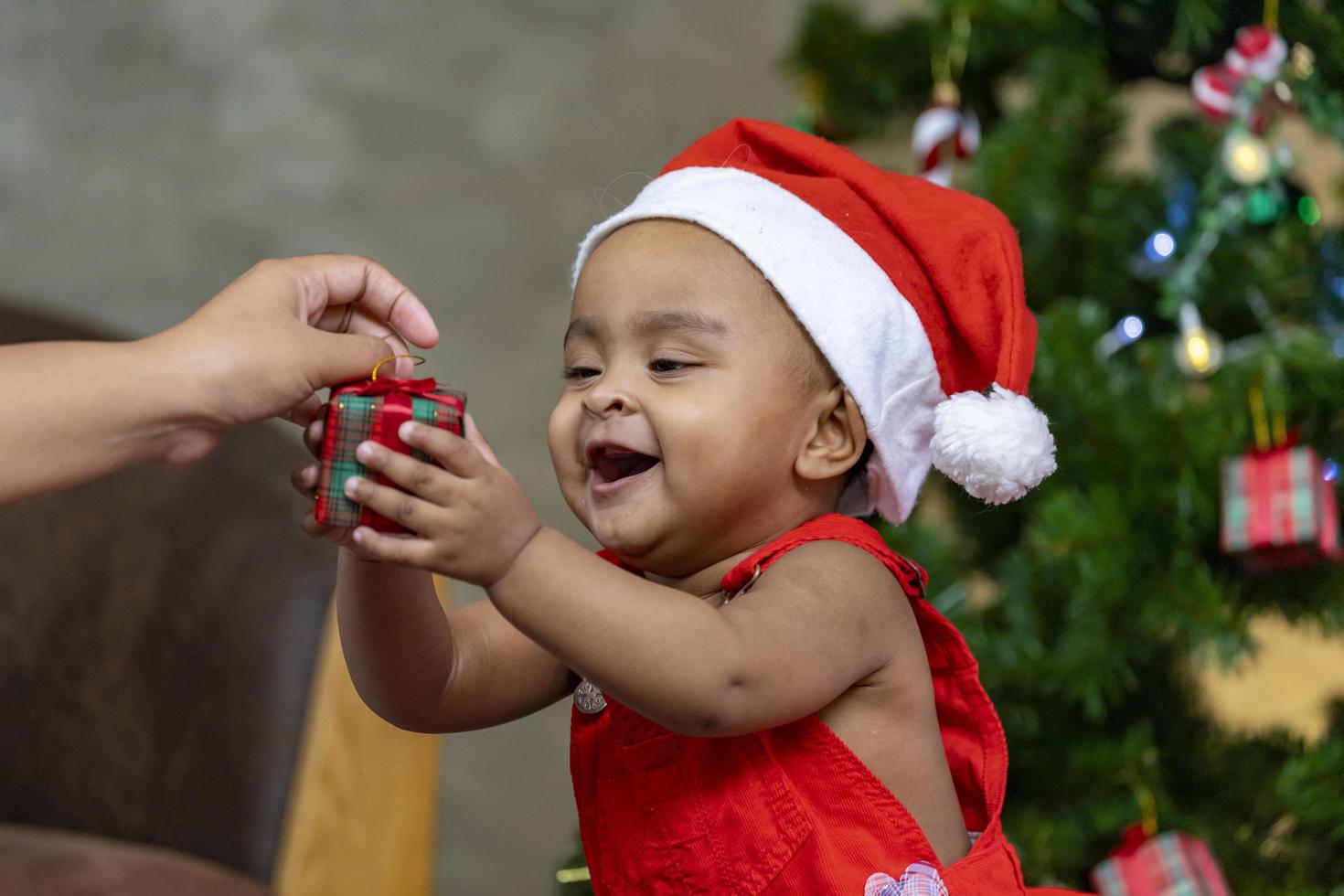 el bebé afroamericano sonríe alegremente mientras recibe una pequeña caja de regalo de los padres mientras se viste con un vestido de navidad y un sombrero de santa con un árbol de navidad en la parte posterior para el concepto de celebración de temporada foto