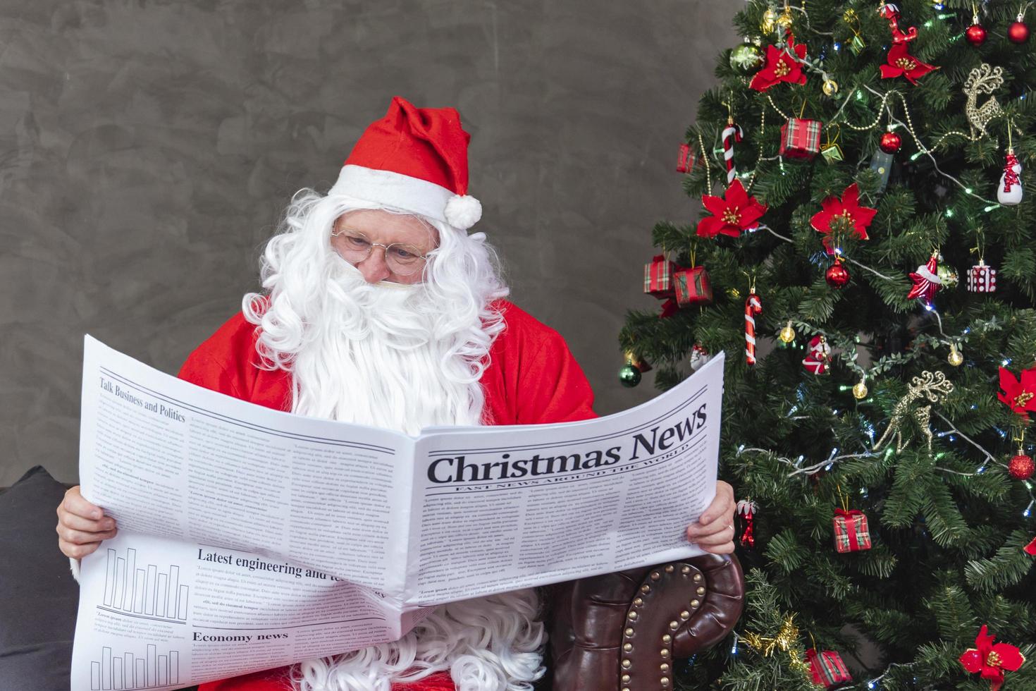 santa claus está leyendo noticias navideñas del periódico mientras está sentado en el sofá junto al árbol de navidad para una situación económica de vacaciones largas e información sobre el informe de transporte de accidentes de tráfico foto