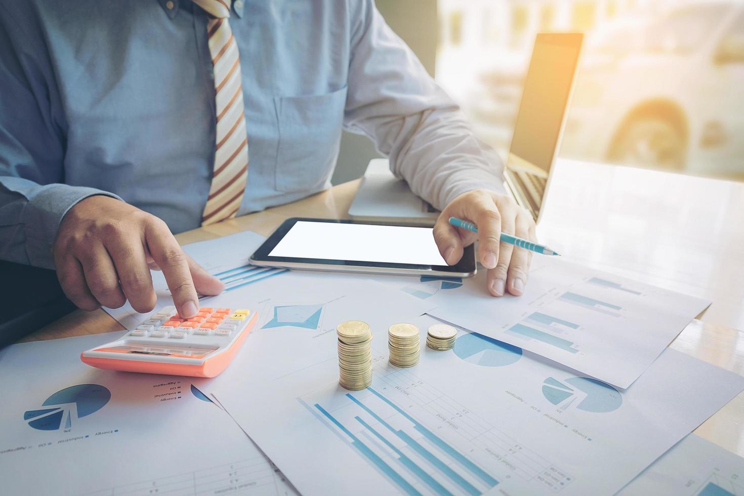 businessman using calculator and tablet with stacked coins arranged at office desk and many document data graph in morning light, business concept. photo