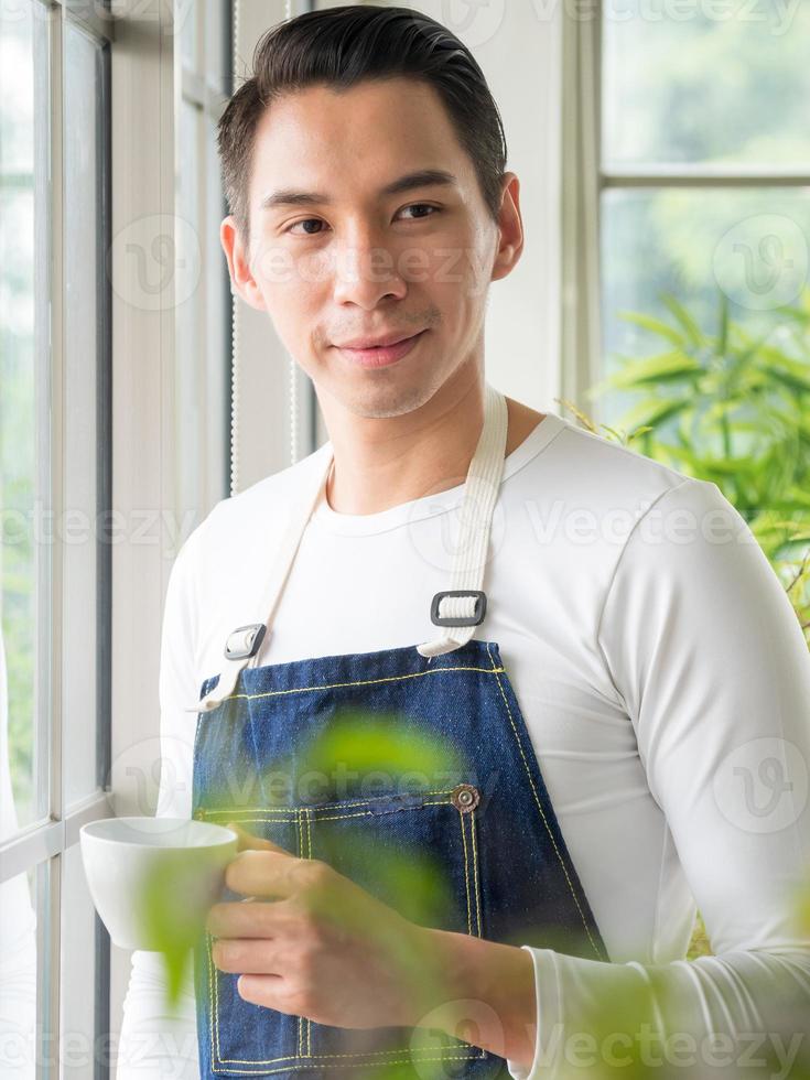 jardinero adulto joven asiático una persona guapo de pie piensa mira y sonríe relajándose feliz bebiendo una taza de café en la cafetería de la habitación con ventana de vidrio y árboles verdes en la mañana de primavera. foto