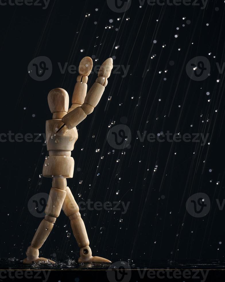 Wooden mannequin of a man in the rain pulls his hands up on a black background. The concept of joy. A wooden figure walks through puddles. photo