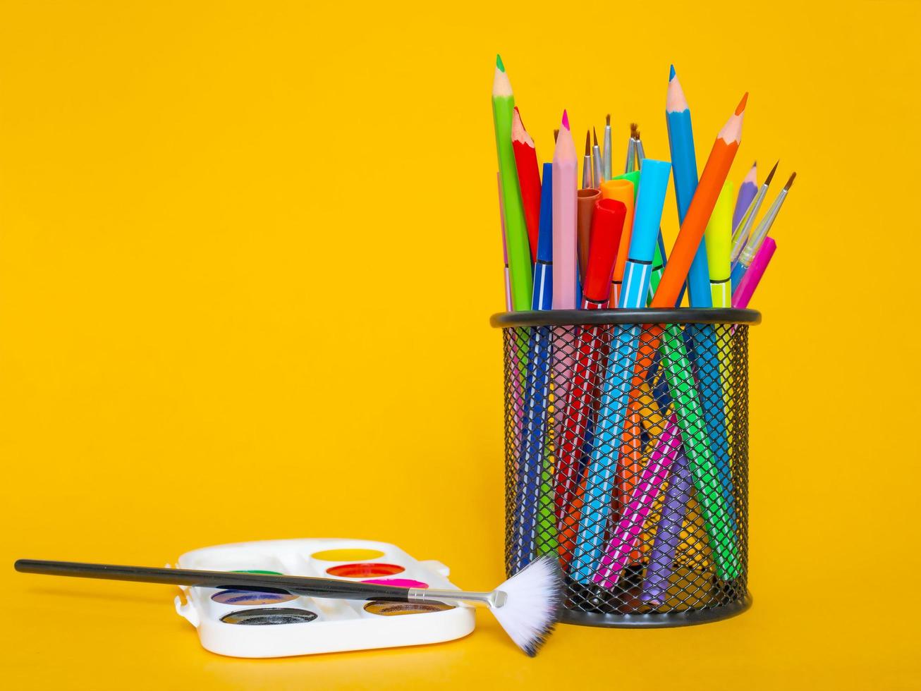 Colored pencils in a pencil case on a yellow background with a white sheet of paper and paints photo