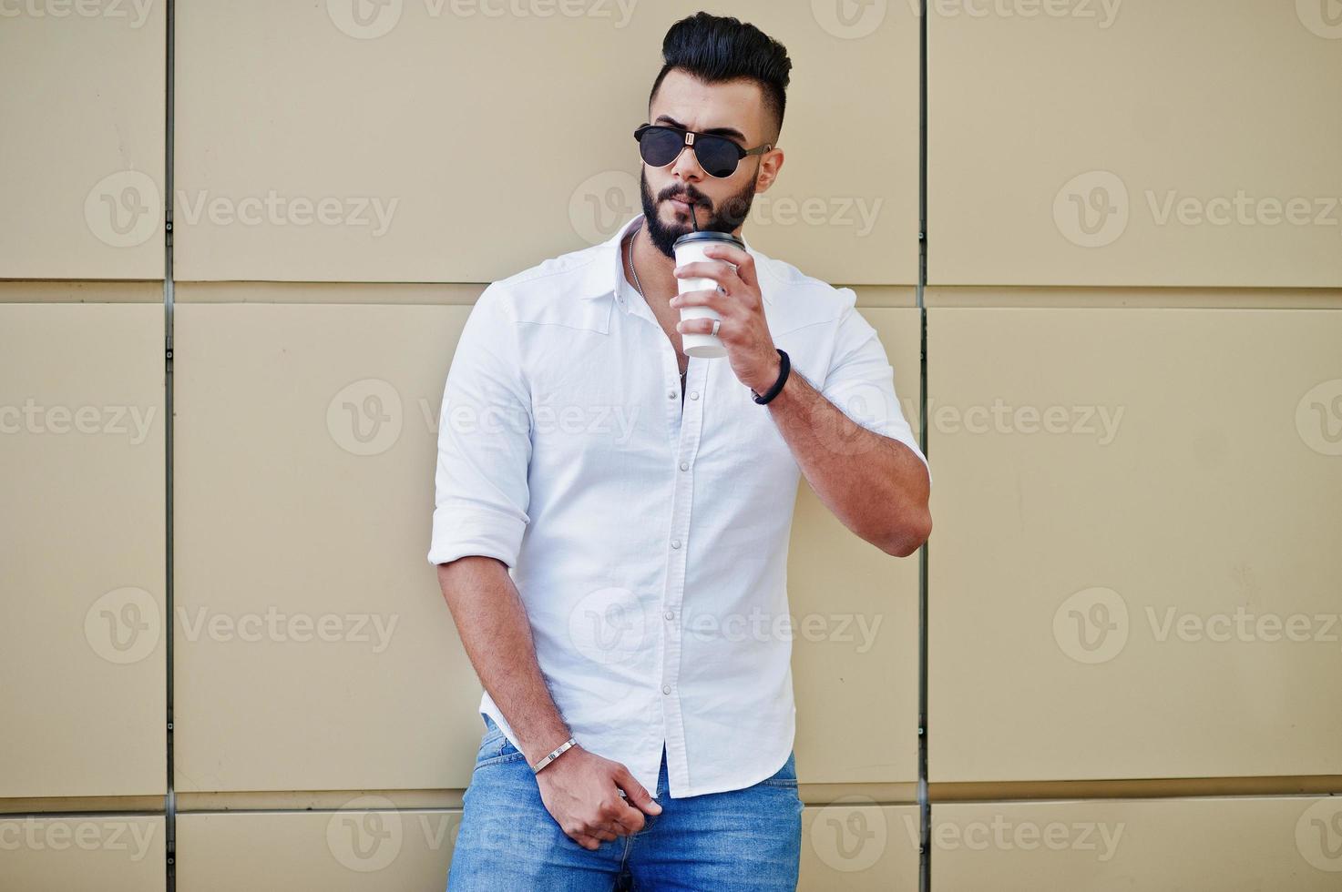 Elegante modelo de hombre árabe alto con camisa blanca, jeans y gafas de sol en la calle de la ciudad. Barba atractivo chico árabe con taza tomando café contra la pared dorada. foto