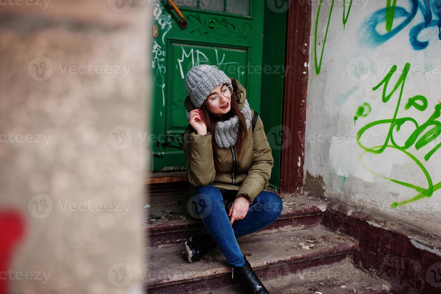 Portrait of brunette girl in gray scarf and hat, glasses sitting against urban entrance. photo