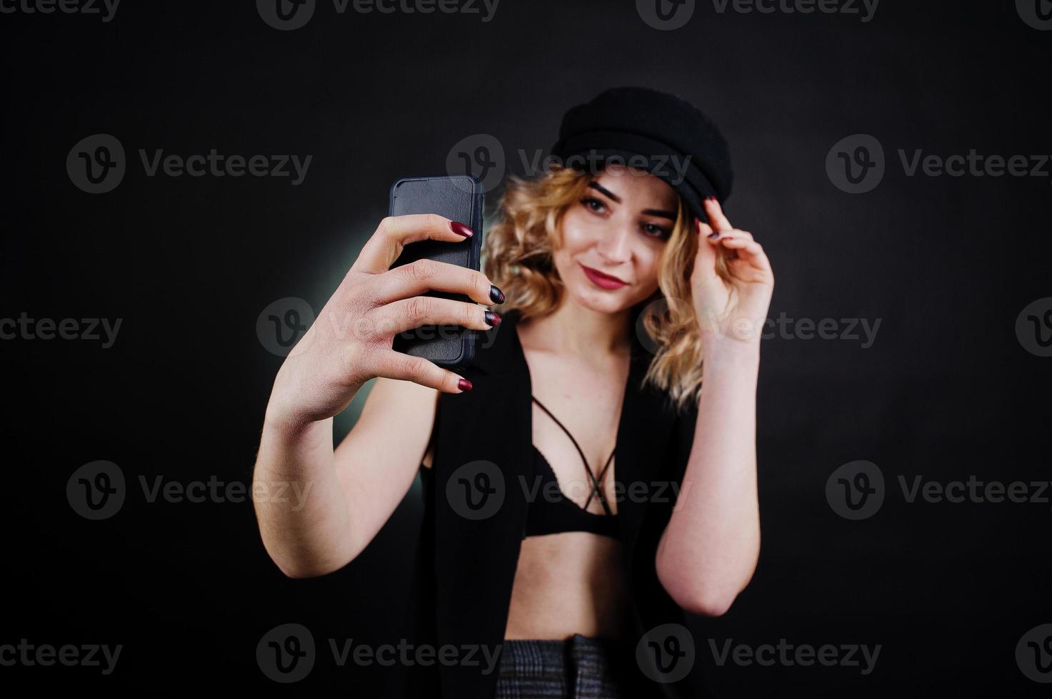 Studio portrait of blonde girl in black wear, bra and cap making selfie against dark background. photo