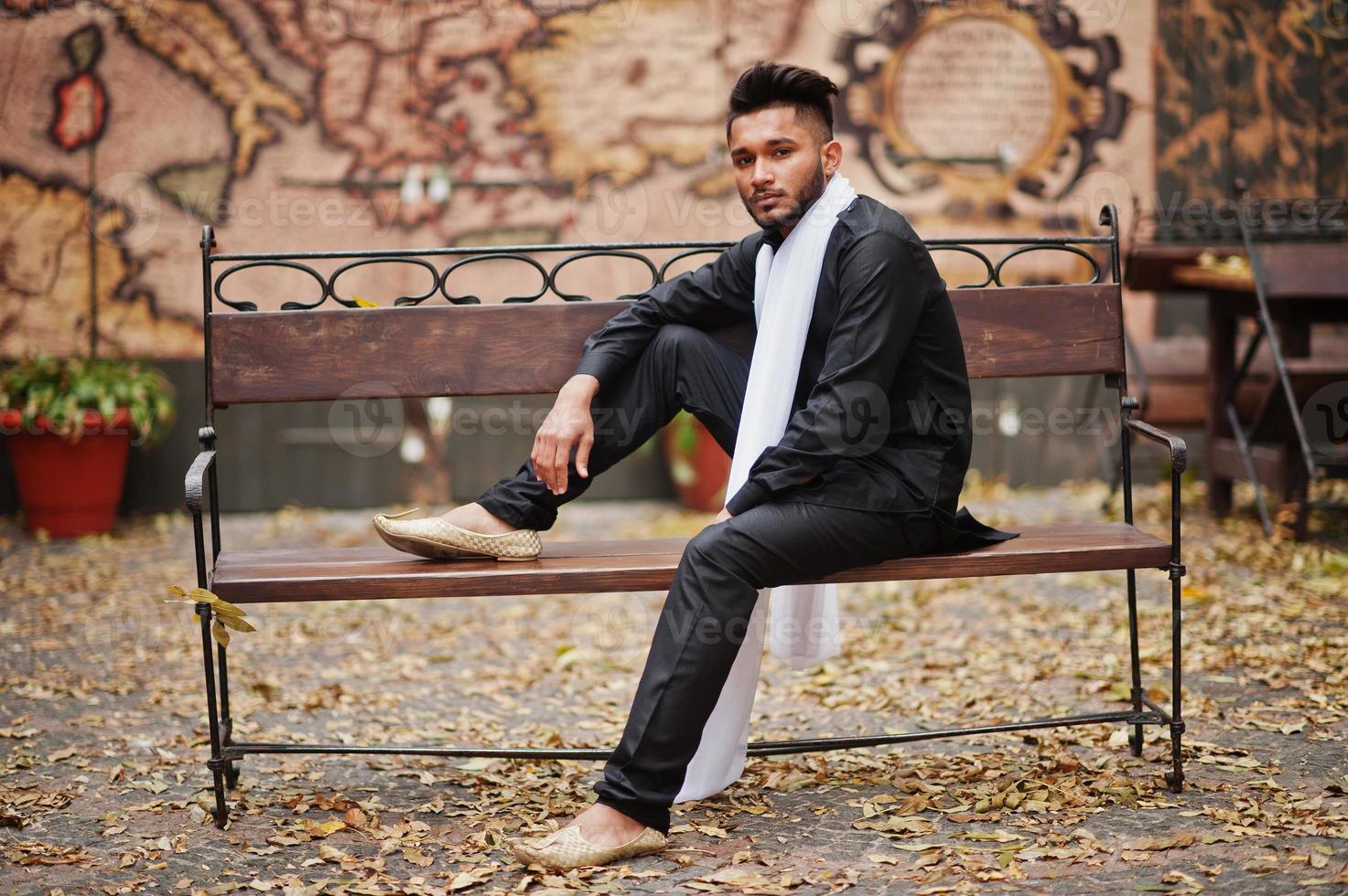 hombre indio elegante vestido con ropa tradicional negra con bufanda blanca sentado al aire libre en un banco. foto