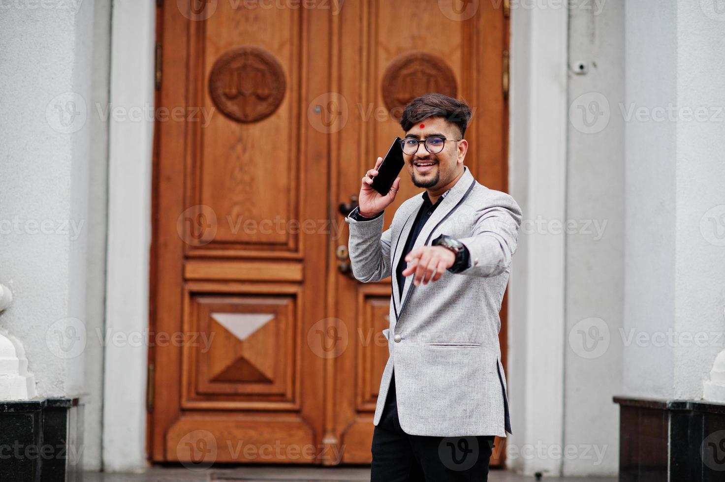 hombre indio elegante con bindi en la frente y anteojos, vestido con traje gris posado al aire libre contra la puerta del edificio, hablando por teléfono móvil y mostrando el dedo a la cámara. foto