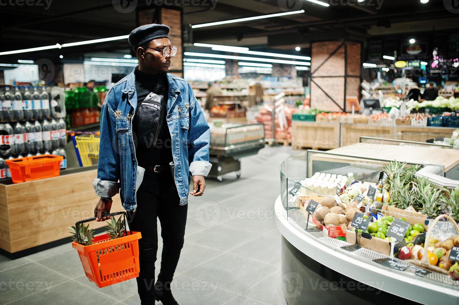 Elegante hombre afroamericano casual con chaqueta de jeans y boina negra sosteniendo una canasta con piñas en la sección orgánica de frutas del supermercado. foto