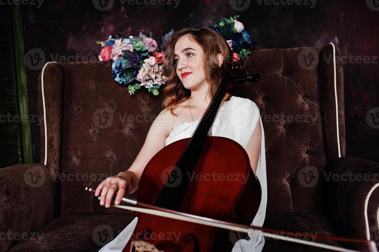 Pretty young gilrl musician in white dress with double bass sitting on brown vintage sofa. photo