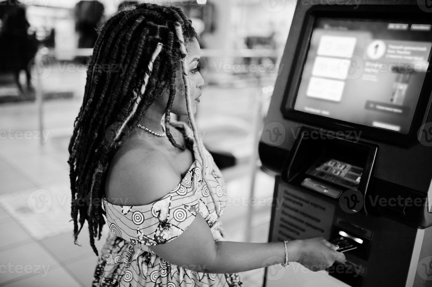 linda chica afroamericana de pequeña altura con rastas, vestida con un vestido amarillo de color, contra un cajero automático con tarjeta de crédito a mano. en blanco y negro. foto