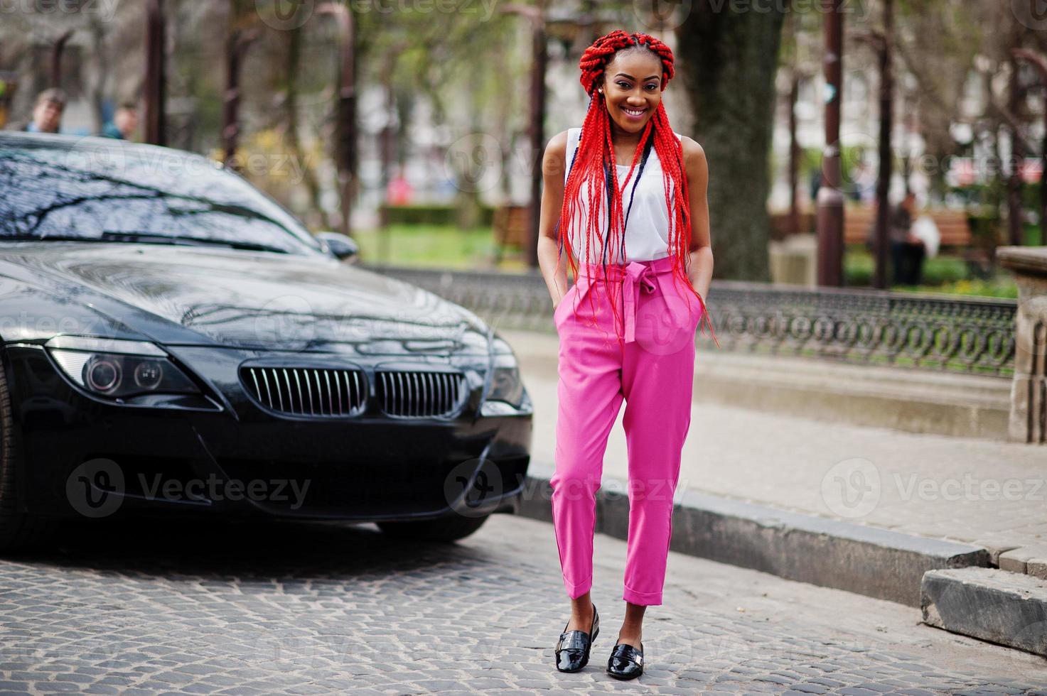 Fashionable african american girl at pink pants and red dreads posed outdoor against black car. photo