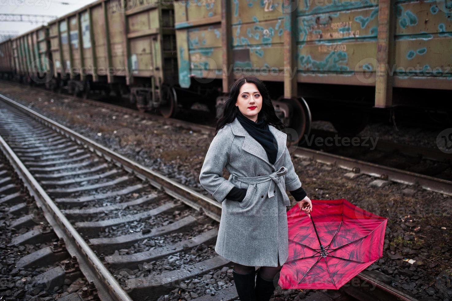 Brunette girl in gray coat with red umbrella in railway station. photo