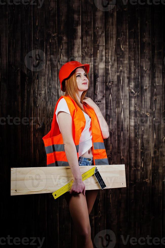 Engineer woman in orange protect helmet and building jacket against wooden background holding board and ruler. photo