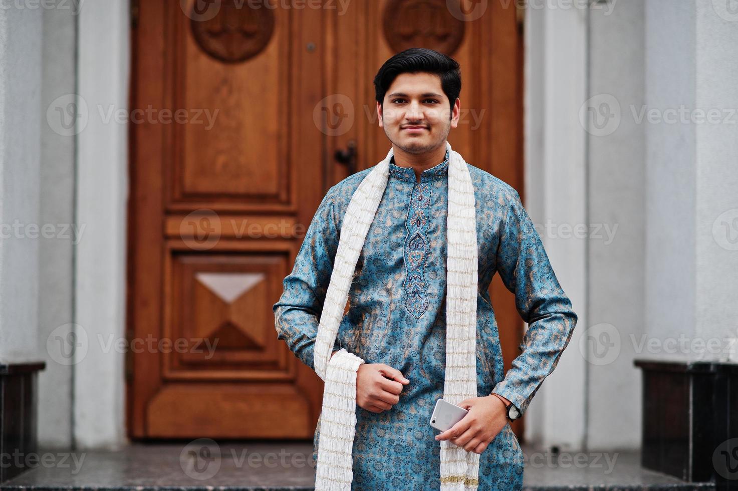 Indian man wear on traditional clothes with white scarf posed outdoor against door of building with mobile phone at  hand. photo