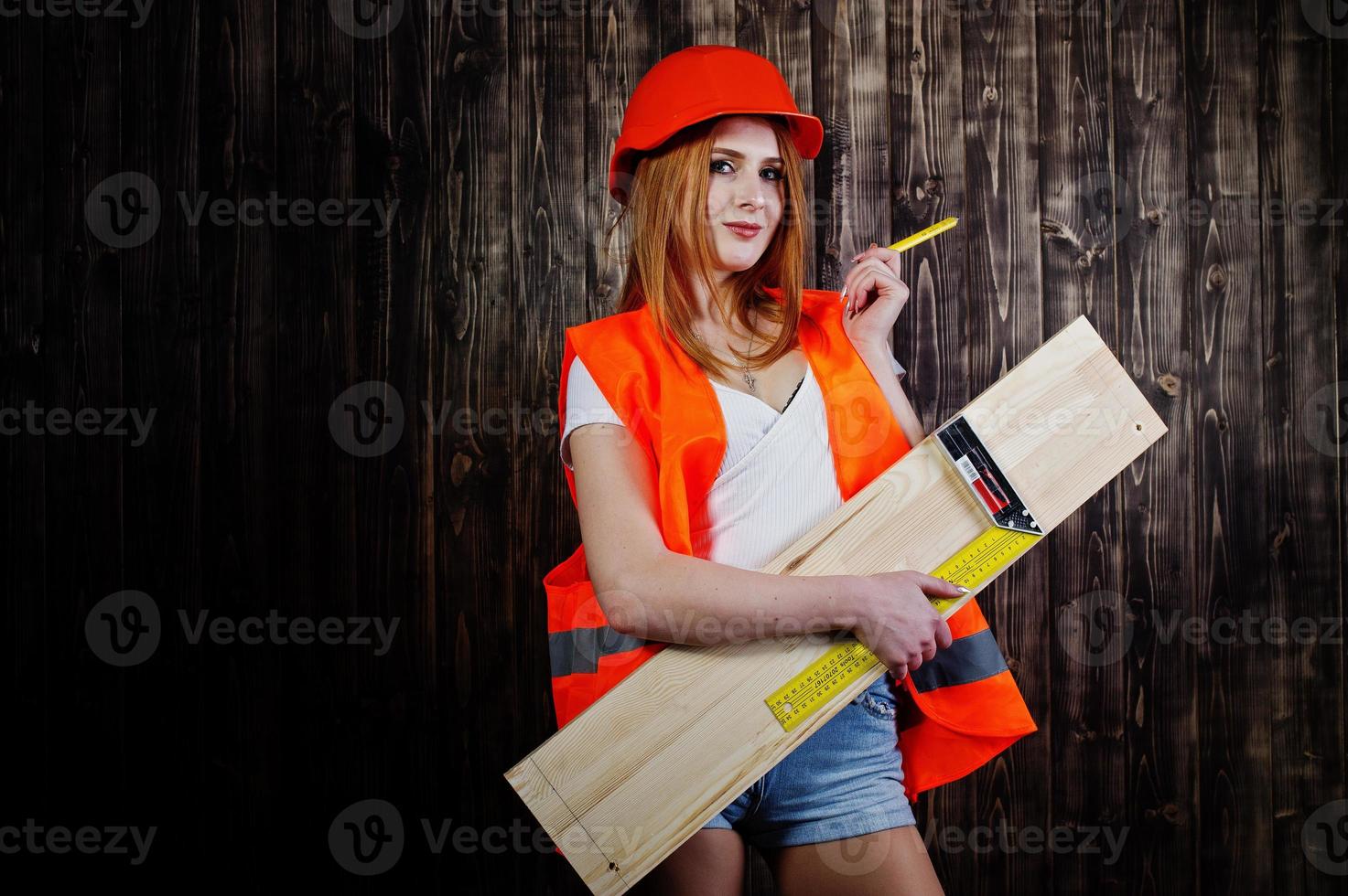 Engineer woman in orange protect helmet and building jacket against wooden background holding board and ruler. photo