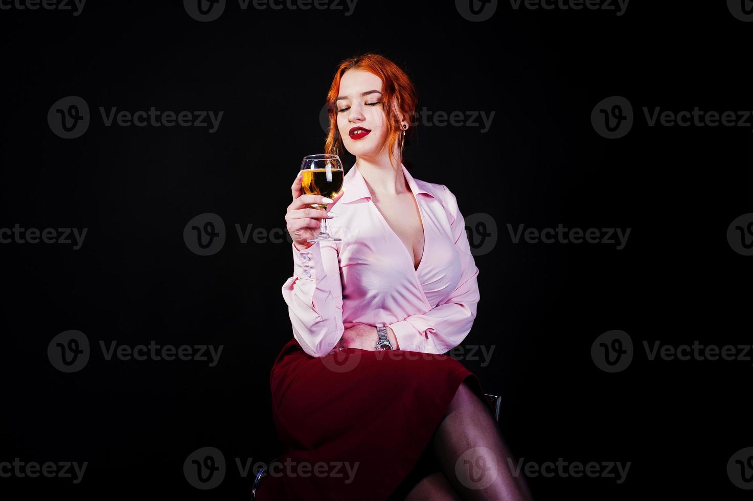 Gorgeous red haired girl in pink blouse and red skirt with glass of wine at hand isolated on black. photo