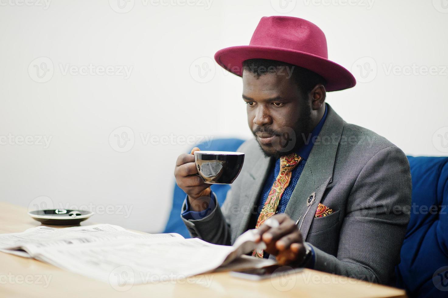 Stylish African American man model in gray jacket tie and red hat drink coffee at cafe and read newspapers. photo