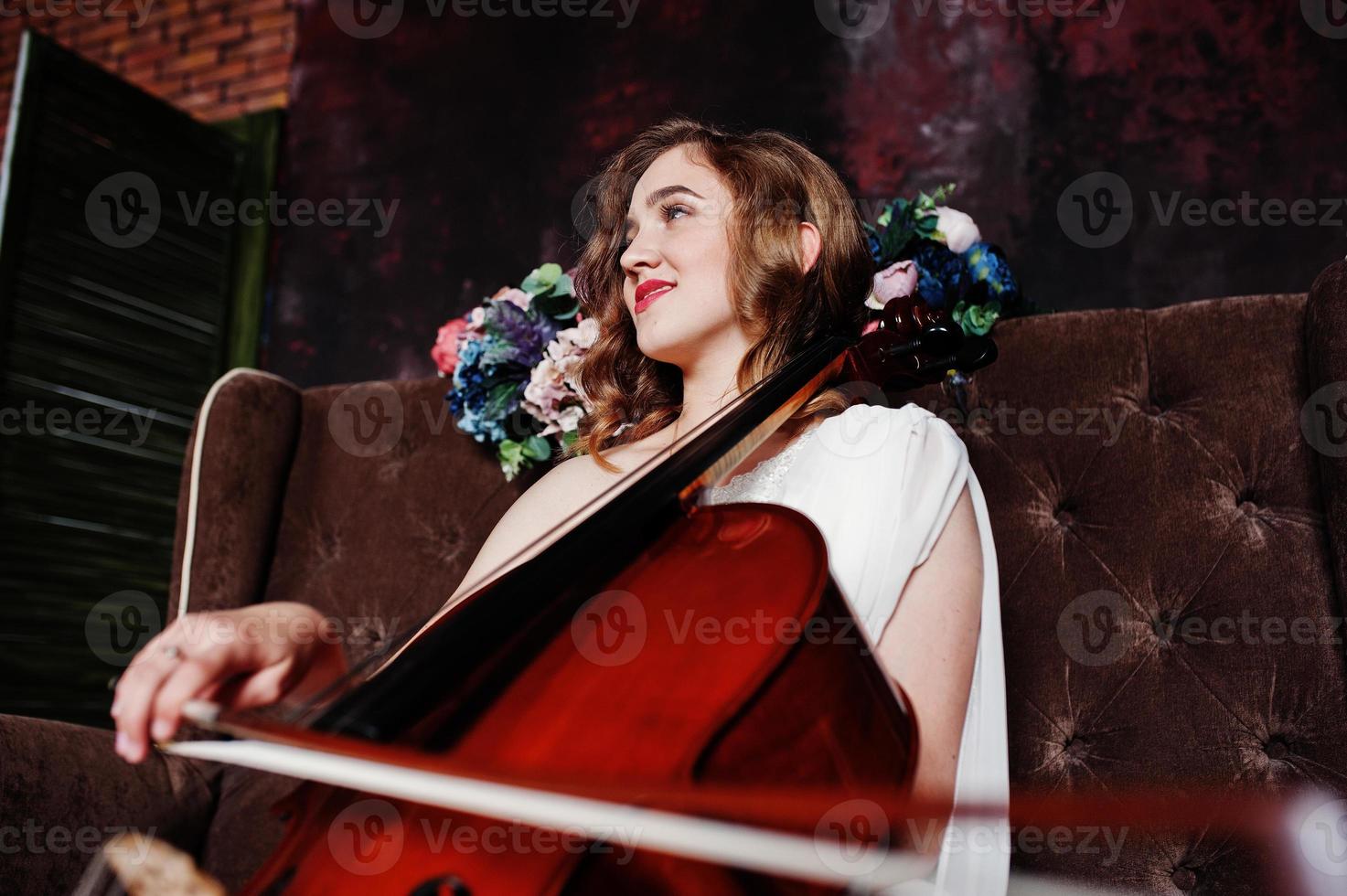 Pretty young gilrl musician in white dress with double bass sitting on brown vintage sofa. photo