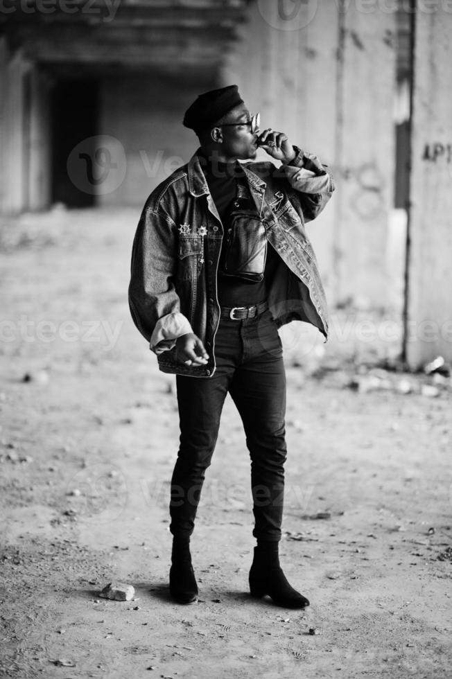African american man in jeans jacket, beret and eyeglasses, smoking cigar at abandoned factory. photo