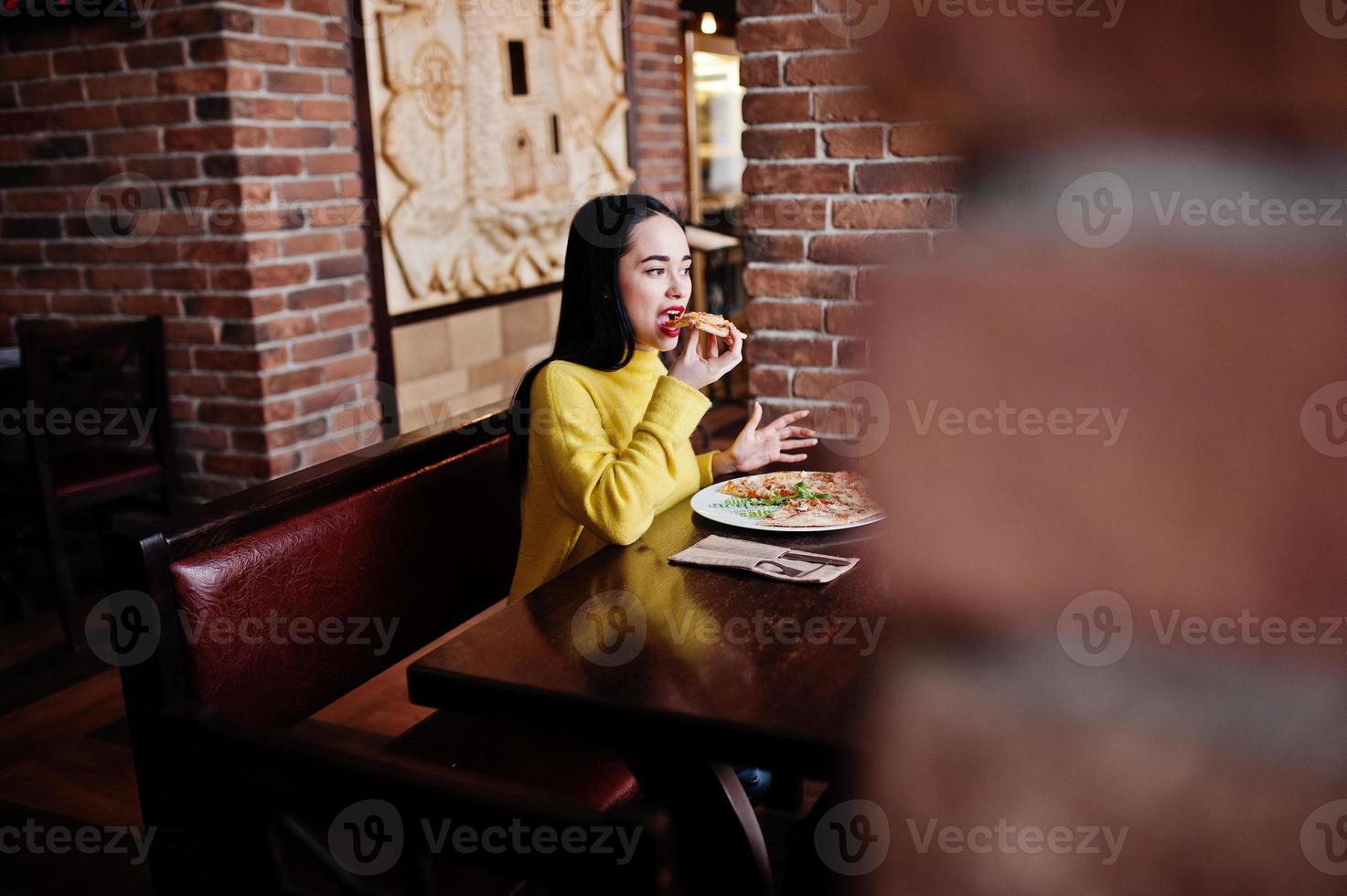 Funny brunette girl in yellow sweater eating pizza at restaurant. photo