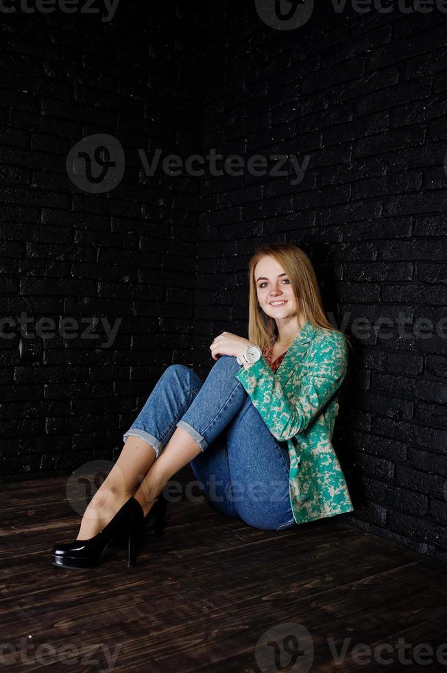 Stylish blonde girl in jacket and jeans against brick black wall at studio. photo