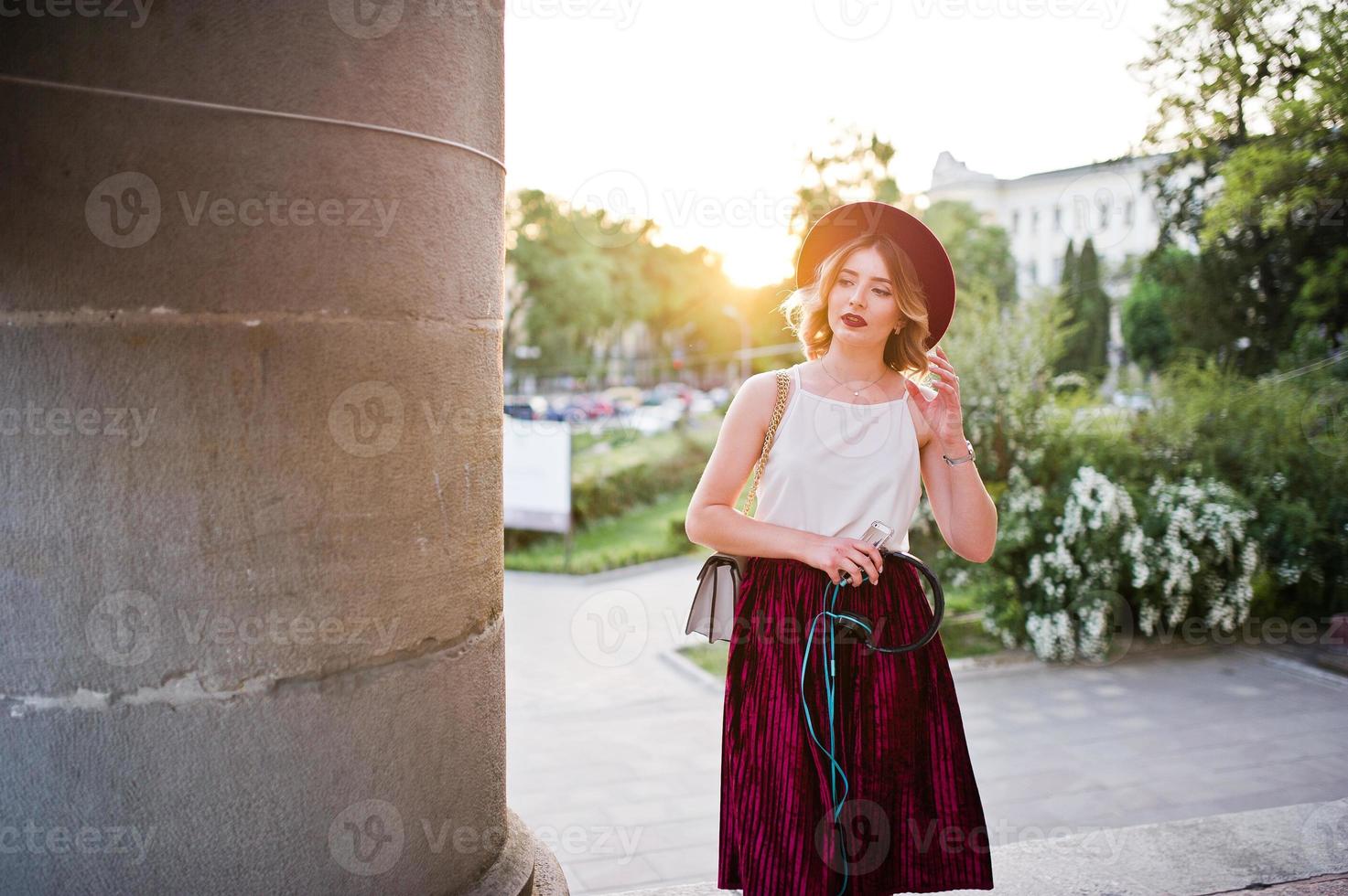 Fashionable and beautiful blonde model girl in stylish red velvet velour skirt, white blouse and hat, posed with phone and earphones against column at sunset. photo