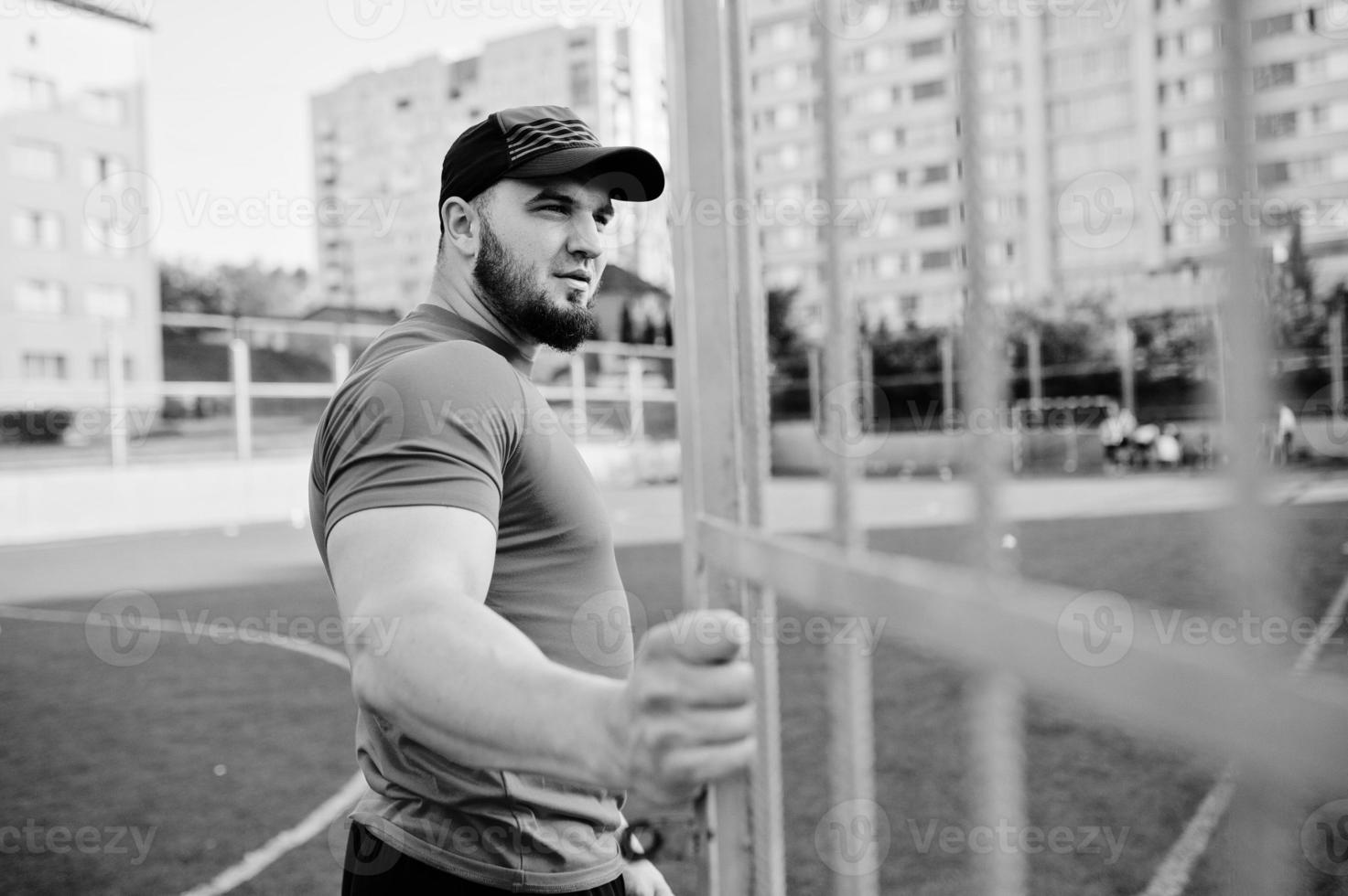 Young brutal bearded muscular man wear on red shirt, shorts and cap at stadium. photo