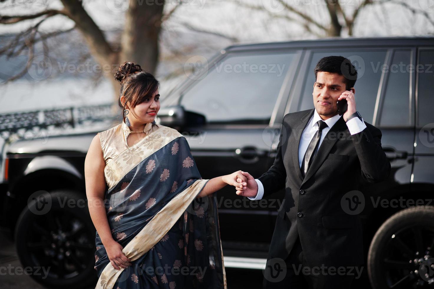 elegante y de moda amigos indios pareja de mujer en sari y hombre en traje posaron contra un rico camión suv negro. tipo hablando por teléfono móvil. foto