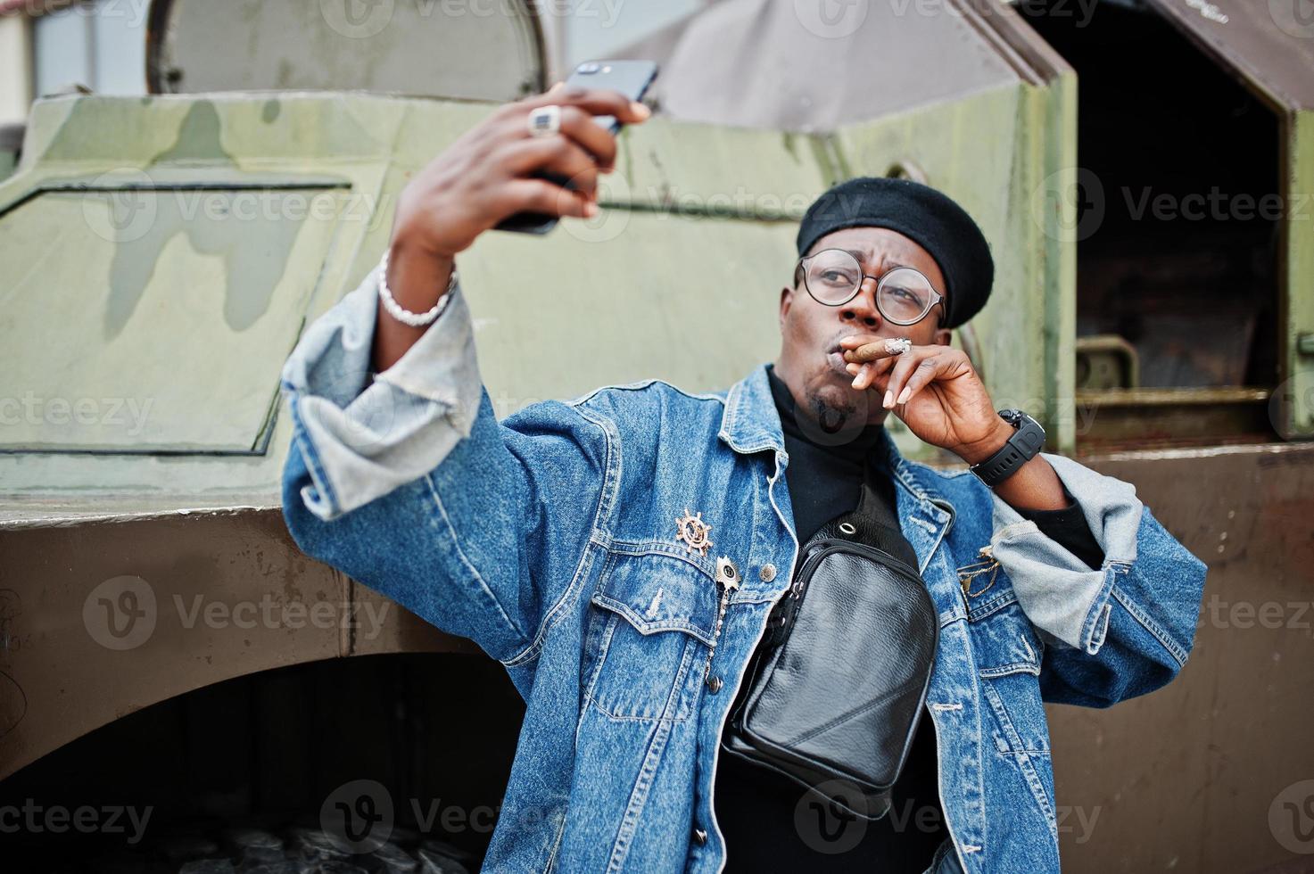 hombre afroamericano con chaqueta de jeans, boina y anteojos, fumando cigarro y posó contra un vehículo blindado militar btr, haciendo selfie por teléfono. foto